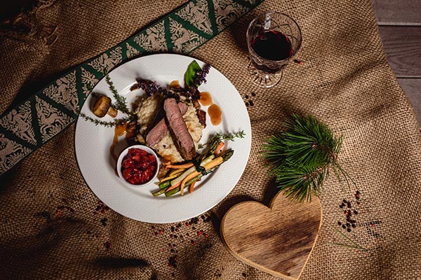 Blick auf einen schön angerichteten Teller mit tranchiertem Fleisch, Risotto und Gemüse, daneben Rotwein und ein Holzherz.