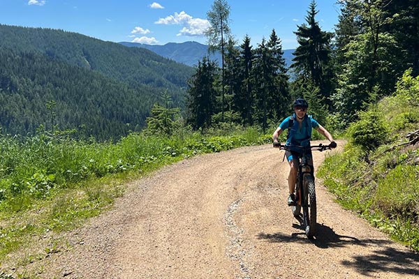 Eine Mountainbikerin fährt die Forststraße hinauf - im Hintergrund wundervoller Blick auf die bewaldeten Berge.