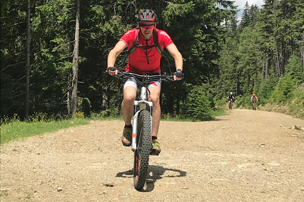 Das Bild zeigt einen Mountainbiker, der auf einem breiten, unbefestigten Waldweg fährt. Er trägt einen Helm, ein Trikot und eine kurze Hose. Im Hintergrund sind zwei weitere Radfahrer zu sehen, die ebenfalls auf dem Weg unterwegs sind. Hohe, grüne Bäume säumen den Weg und verleihen der Szene eine natürliche, ruhige Atmosphäre.