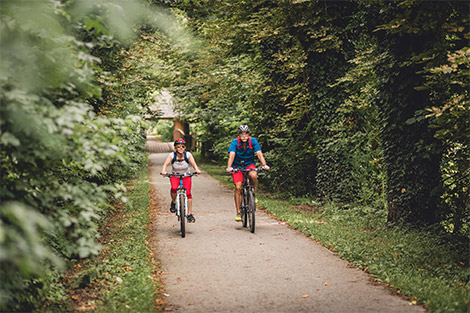 Zwei Pensionisten sind mit dem Fahrrad auf dem Mürtzalradweg unterwegs - links und rechts Sträucher und Laubbäume